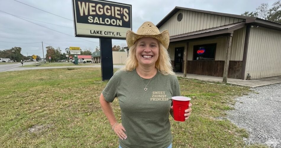 Smiling blonde woman in a straw cowboy hat and green t-shirt holding a red solo cup in front of Weggie's Saloon sign in Lake City, FL.