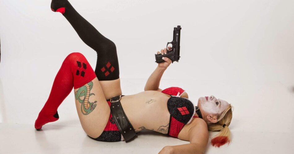 Harley Quinn Cosplay by Queen Daisy Chain. She's laying on her back holding a prop gun. This black and red Harley Quinn is inspired by Batman the Animated Series.