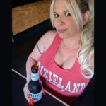 Queen Daisy Chain holding a beer at the bar. She's wearing a red tank top that says "Dixieland Delight"
