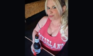 Queen Daisy Chain holding a beer at the bar. She's wearing a red tank top that says "Dixieland Delight"