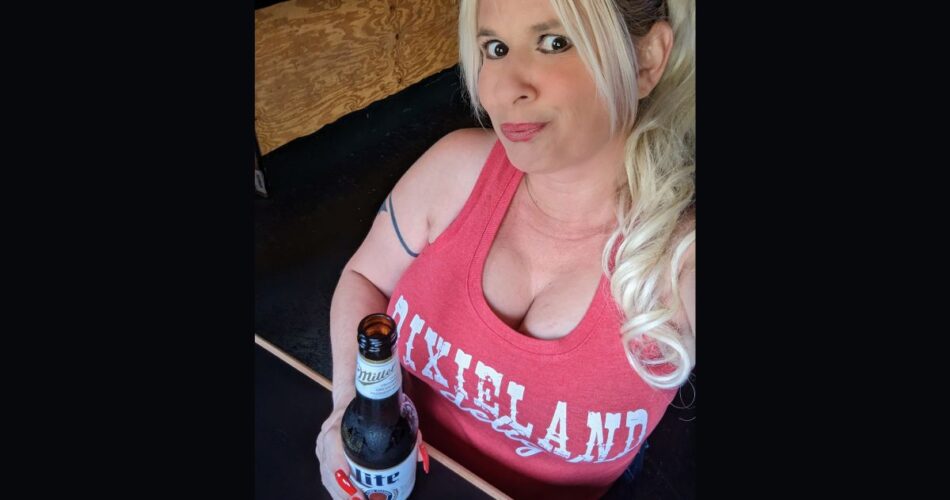 Queen Daisy Chain holding a beer at the bar. She's wearing a red tank top that says "Dixieland Delight"